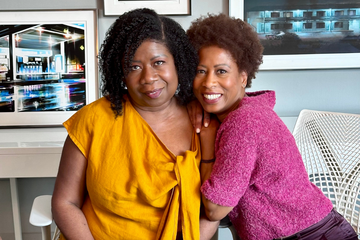 Coils to Locs Co-founders Dianne (Gold Blouse) and Pamela (pink cropped sweater) posing, seated, in an office space.